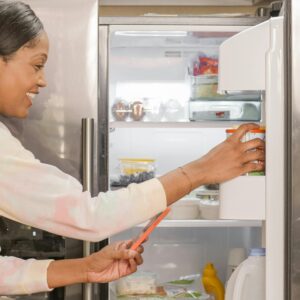 Smiling woman using mobile phone while organizing refrigerator.